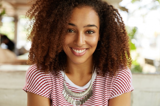 Portrait de femme heureuse à la peau foncée avec une coiffure afro touffue bouclée, repose dans le café