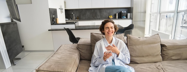 Photo gratuite portrait d'une femme heureuse qui rit d'une vidéo drôle sur son téléphone tenant un smartphone assise sur le canapé à