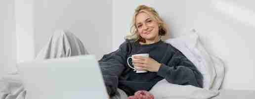 Photo gratuite portrait d'une femme heureuse en train de profiter d'un jour de congé à la maison allongée dans son lit en regardant une émission de télévision sur un ordinateur portable.