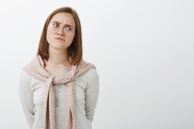 Portrait de femme intelligente sombre perplexe dans des verres avec de courts cheveux bruns se détournant à droite en fronçant les sourcils et en pinçant les lèvres étant concerné et réfléchissant à résoudre le dilemme sur mur gris