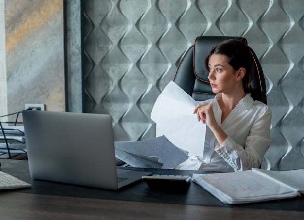 Portrait de femme jeune employé de bureau assis au bureau avec des documents et un ordinateur portable à la recherche de côté avec une expression pensive pensant travailler au bureau