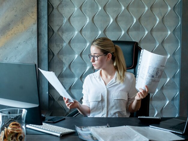 Portrait de femme jeune employé de bureau assis au bureau avec des documents les regardant avec une expression de confiance grave sur le visage travaillant au bureau