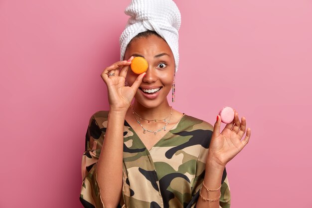 Portrait de femme joyeuse et animée joue avec de délicieux macarons, habillé avec désinvolture, porte une serviette enveloppée sur la tête, aime manger un dessert français, pose contre un mur rose. Les gens et la nourriture