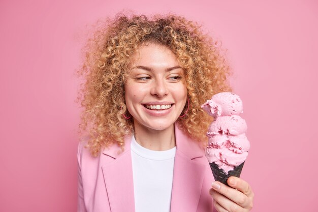 Le portrait d'une femme joyeuse aux cheveux bouclés sourit largement regarde une glace appétissante dans une gaufre noire mange un délicieux dessert d'été porte une tenue élégante sent la tentation isolée sur un mur rose