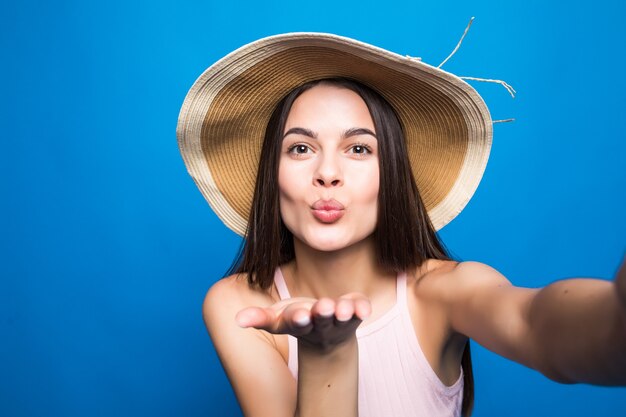 Portrait de femme joyeuse envoyer des baisers aériens en robe et chapeau d'été en prenant un selfie isolé sur un mur bleu.