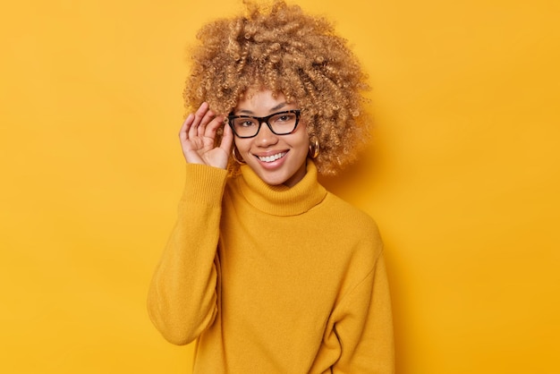 Portrait d'une femme joyeuse garde la main sur le bord des lunettes sourit joyeusement porte un pull décontracté se sent positif isolé sur fond jaune vif se tient heureux à l'intérieur. Les gens et les émotions