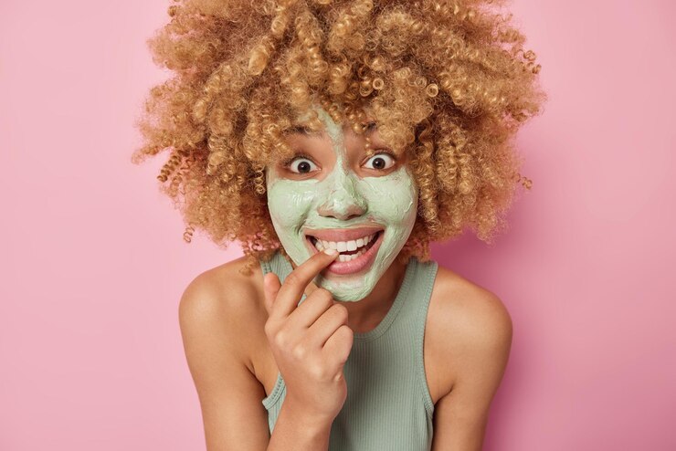Photo gratuit portrait d'une femme joyeuse surprise garde le doigt dans la bouche a une expression heureuse applique un masque de beauté pour le rajeunissement entend de bonnes nouvelles porte un t-shirt se tient les épaules nues isolé sur un mur rose