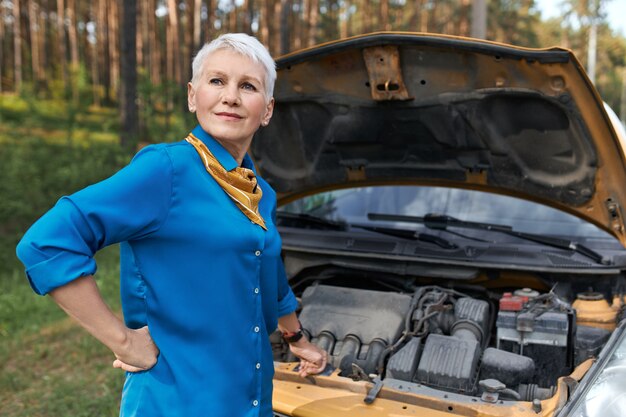 Portrait de femme mature aux cheveux courts blonds ayant une expression faciale frustrée parce que la voiture est cassée. Femme d'âge moyen stressé en attente de service après une panne de véhicule, ouverture du capot