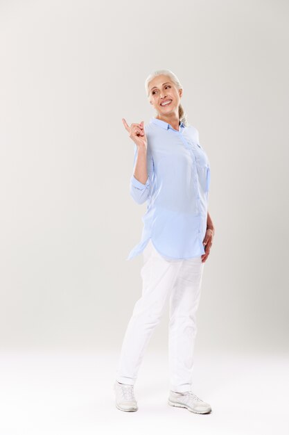 Portrait de femme mature ludique en chemise bleue et pantalon blanc, debout et pointant avec le doigt, regardant de côté