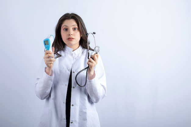 Portrait de femme médecin tenant un thermomètre et un stéthoscope sur un mur blanc.