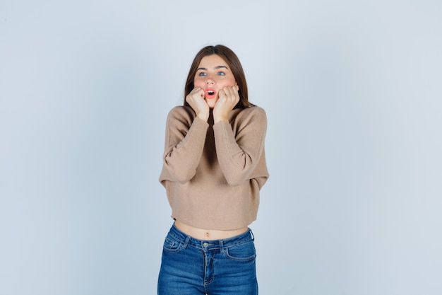 Portrait d'une femme merveilleuse gardant les poings sur les joues tout en ouvrant la bouche en pull, en jean et en regardant la vue de face choquée