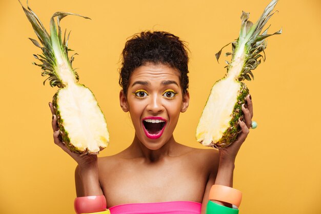 Portrait de femme métisse ravie avec apparence de mode tenant deux moitiés d'ananas dans les deux mains isolées, sur le mur jaune