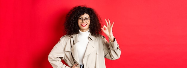 Photo gratuite portrait d'une femme à la mode confiante dans des verres et un trench-coat montrant un geste correct pour approuver ou agr