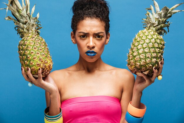 Portrait de femme mulâtre sérieuse avec une apparence élégante face fronçant les sourcils étant bouleversé tout en tenant deux ananas mûrs dans les deux mains isolées, sur le mur bleu