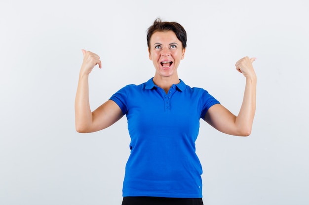 Portrait de femme mûre pointant vers l'arrière avec les pouces en t-shirt bleu et à la vue de face confiante