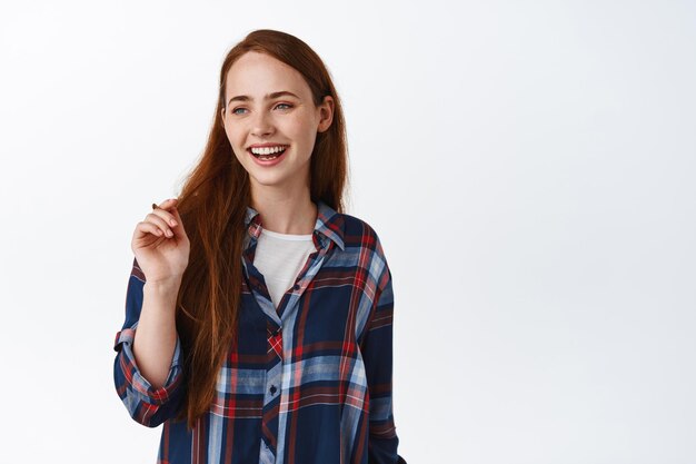 Portrait d'une femme naturelle souriante aux cheveux longs rouges, riant et ayant l'air heureux, regardant de côté le logo, debout dans une chemise à carreaux de vêtements décontractés, fond blanc