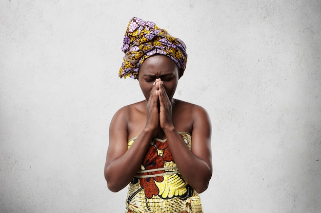 Portrait De Femme Noire Mendiant En Vêtements Traditionnels En Appuyant Sur Ses Paumes Ensemble Fermant Les Yeux Implorant La Bonne Chance De Ses Enfants. Femme Au Foyer Africaine Religieuse Priant Pour Le Bien-être De La Famille