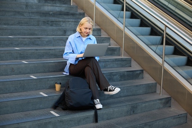 Photo gratuite portrait d'une femme avec un ordinateur portable trouvé point wifi assis sur les escaliers à l'extérieur buvant du café travaillant sur