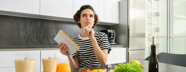 Photo gratuite portrait d'une femme pensante avec un cahier de cuisine écrivant les ingrédients de la recette décidant d'un repas