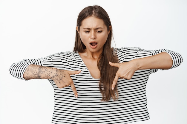 Photo gratuite portrait de femme pointant les doigts vers le bas avec un visage choqué. modèle féminin avec des tatouages et des piercings à la recherche d'onwhelmed et frustré sur blanc.