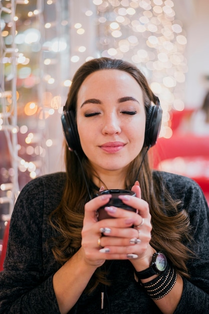 Portrait de femme portant des écouteurs tenant la tasse près des lumières de Noël