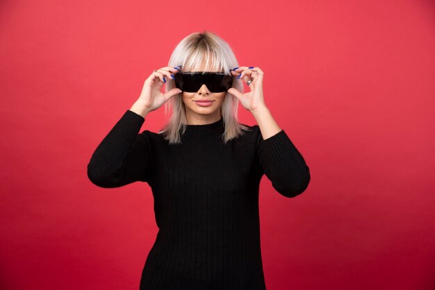 Portrait de femme portant des lunettes noires sur un mur rouge.