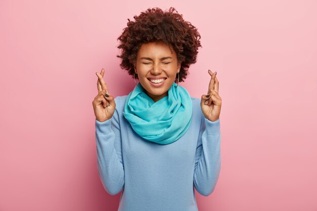 Portrait de femme ravie aux cheveux afro, garde les doigts croisés, croit en la bonne chance, sourit largement, porte un pull bleu avec écharpe