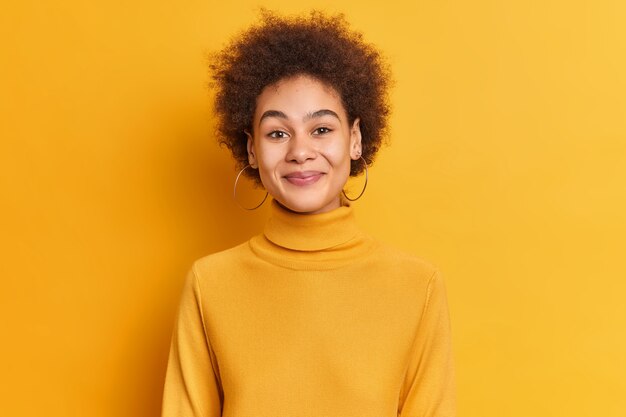 Portrait de femme à la recherche agréable avec des cheveux bouclés étant de bonne humeur sourit satisfait porte col roulé et boucles d'oreilles occasionnels.