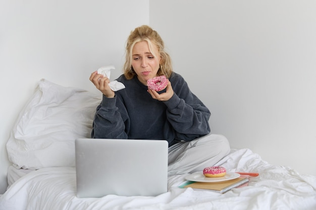 Photo gratuite portrait d'une femme regardant un spectacle triste sur un ordinateur portable mangeant un donut et essuyant ses larmes avec une serviette assise