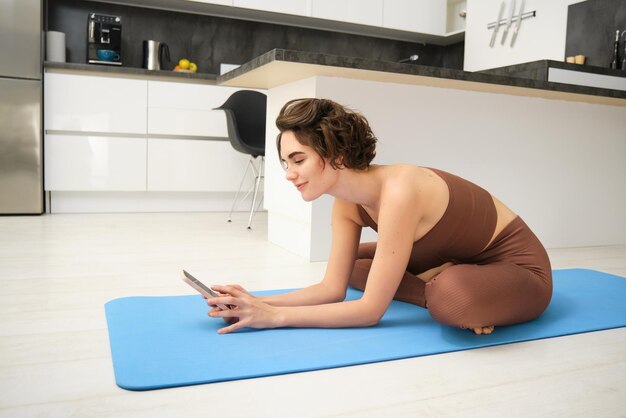 Portrait d'une femme de remise en forme en leggings et soutien-gorge de sport est assise dans la cuisine et fait de l'exercice sur un tapis de yoga