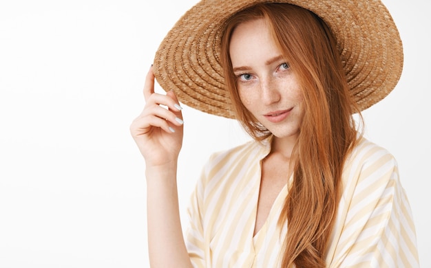 Portrait d'une femme rousse belle mystérieuse et sensuelle élégante souriant flirty regardant avec intérêt et désir toucher chapeau de paille sur la tête posant