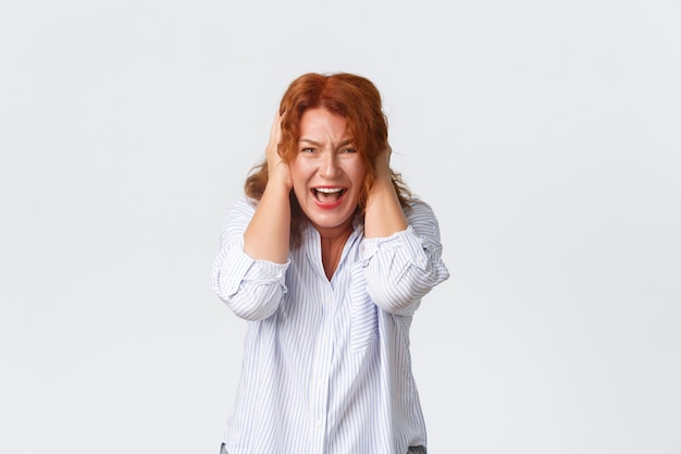 Portrait de femme rousse en détresse et bouleversée en chemise, crier de panique, couvrir les oreilles avec les mains concernées, debout anxieux et insécurisé sur fond blanc. Mère paniquée.