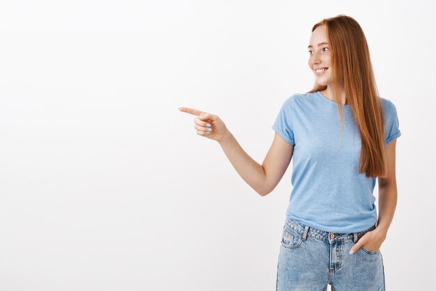 Portrait de femme rousse féminine mignonne et tendre avec des taches de rousseur en t-shirt bleu tenant la main dans la poche pointant et regardant à gauche avec un regard heureux ravi