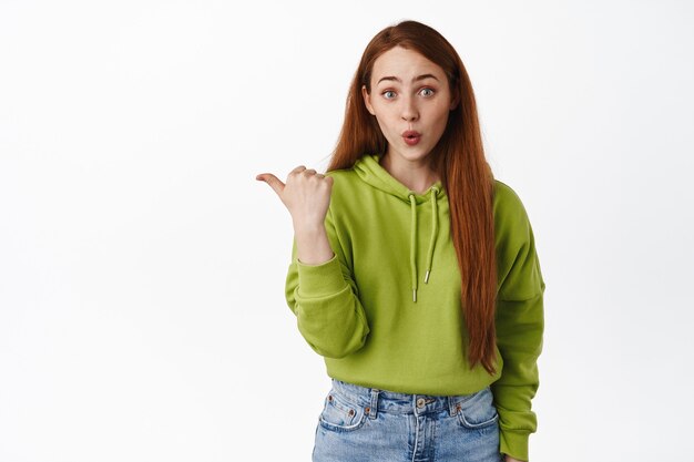 Portrait d'une femme rousse impressionnée, disons wow, pointant le doigt vers la gauche avec un visage étonné sur blanc