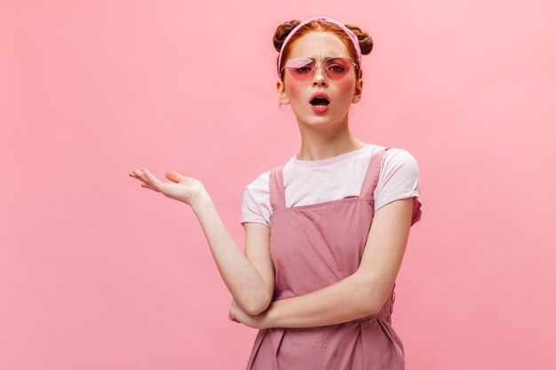 Portrait de femme rousse à lunettes roses et tenue regardant avec indignation la caméra sur fond isolé.