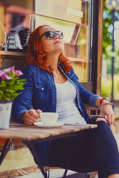Portrait de femme rousse à lunettes de soleil, boit du café dans un café dans une rue.
