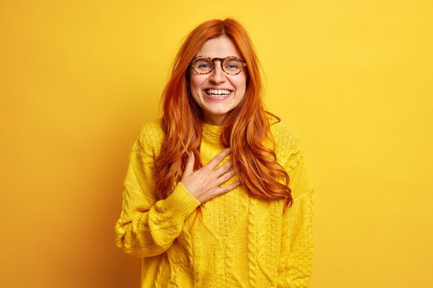 Portrait de femme rousse ravie sourit largement garde la main sur la poitrine sourit exprime largement les bonnes émotions se sent heureux porte des lunettes transparentes chandail décontracté.