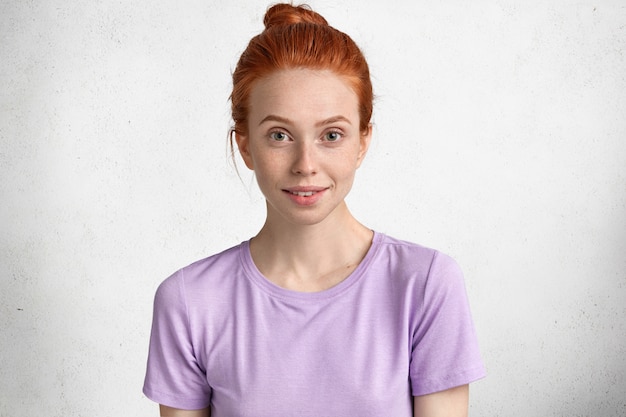 Portrait de femme rousse à la recherche agréable avec noeud de cheveux, regarde joyeusement la caméra, porte un t-shirt violet décontracté, exprime des émotions positives