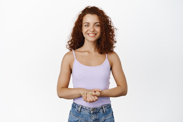 Portrait d'une femme rousse souriante regardant avec une expression de visage amicale et polie, se tenant la main, écoutant et essayant d'aider, aidant, debout sur fond blanc