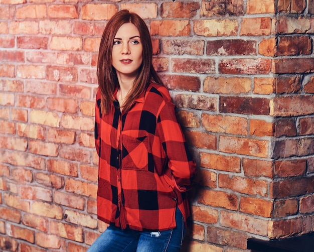 Portrait de femme rousse vêtue d'une chemise polaire rouge et d'un jean sur le mur de briques rouges.