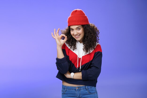 Portrait d'une femme séduisante et affirmée avec un regard confiant et des cheveux bouclés portant un chapeau d'hiver chaud montrant un geste correct et souriant assurant, appréciant et approuvant la tenue impressionnante d'un ami.