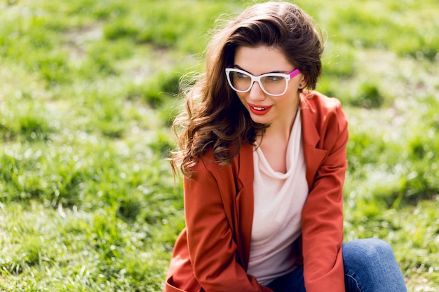 Portrait de femme séduisante avec des lèvres pleines, des lunettes, une veste rouge, une coiffure ondulée assis sur l'herbe verte dans le parc de printemps ensoleillé et souriant