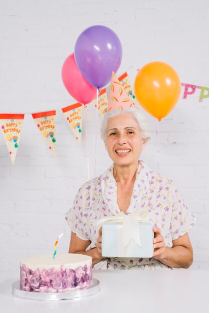 Portrait d&#39;une femme senior heureuse holding cadeau d&#39;anniversaire avec un gâteau sur le bureau