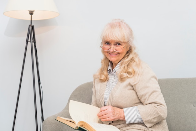 Photo gratuite portrait d'une femme senior souriante assise sur un canapé, tenant le livre à la main