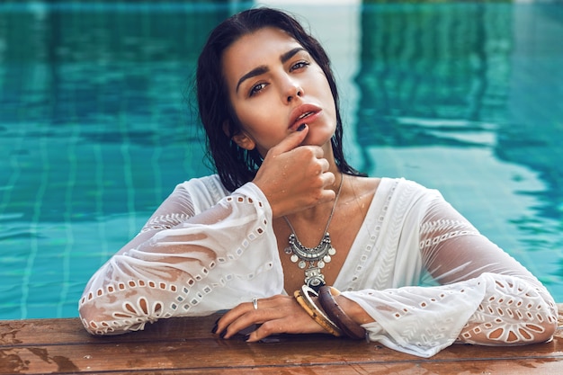 Portrait d'une femme sensuelle incroyable avec un corps bronzé parfait posant dans la piscine, portant des accessoires boho élégants.