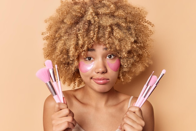 Portrait d'une femme sérieuse aux cheveux bouclés confus tenant des pinceaux cosmétiques pour appliquer le maquillage applique des patchs hydratants pour le rajeunissement de la peau se dresse torse nu isolé sur fond beige.