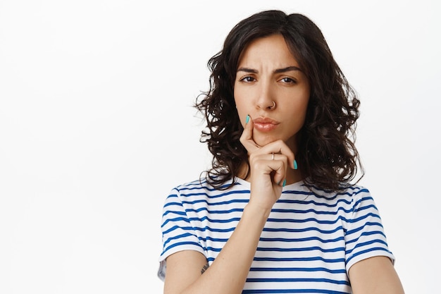 Portrait d'une femme sérieuse réfléchie prend une décision, fronçant les sourcils perplexe, se sentant méfiante et plissant les yeux dans l'incrédulité, dites hmm, debout sur fond blanc