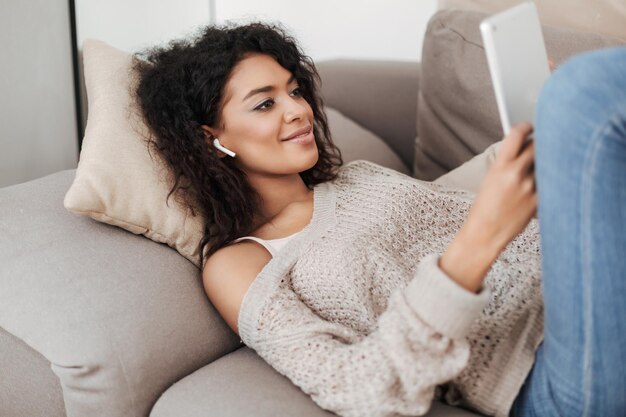 Portrait d'une femme souriante aux cheveux bouclés noirs dans des écouteurs allongés sur un canapé avec une tablette dans les mains à la maison