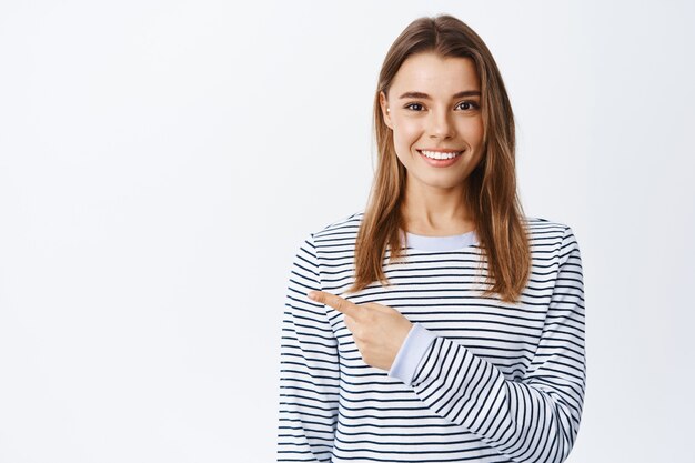 Portrait d'une femme souriante et confiante aux cheveux blonds, pointant le doigt vers la bannière du logo et regardant devant, montrant une publicité sur un mur blanc, portant une chemise à rayures décontractée