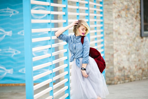 Portrait d'une femme souriante debout avec des cheveux blonds courts, des lèvres rose vif et un maquillage nu appuyé sur une clôture à rayures bleues et blanches